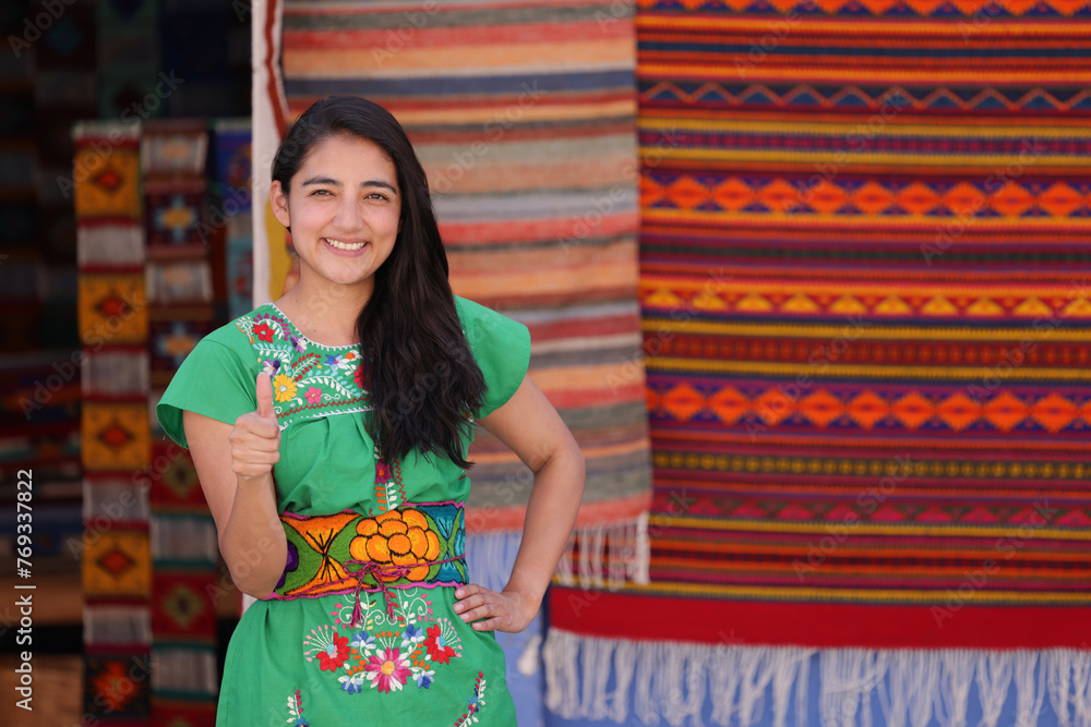 Portrait of smiling young Mexican girl dressed in typical Mexican clothing, looking at the camera with thumb up on a background of Mexican textile crafts.