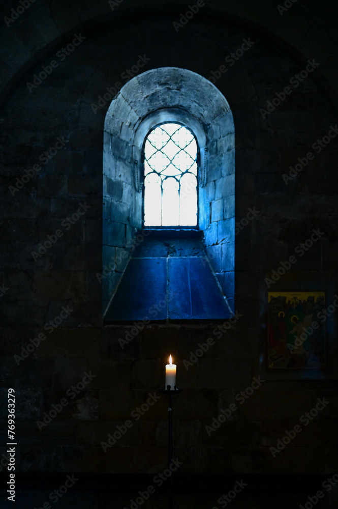 Fototapeta premium A single small, burning candle stands in front of a church window with a mosaic, light shining through the window (3787)