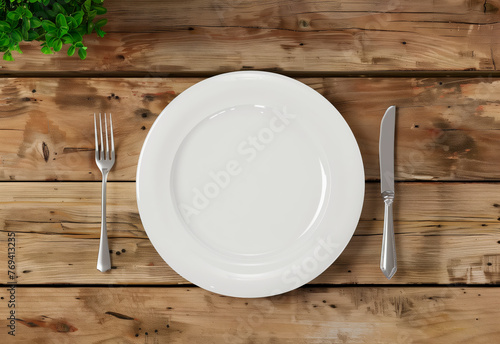 Simple White Plate and Silverware on Wooden Table. A top view of a simple yet elegant dining setup with a pristine white plate flanked by silver fork and knife on a rustic wooden table.