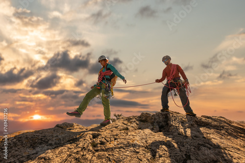 Two people are climbing a mountain together. One of them is wearing a red shirt. The sun is setting in the background