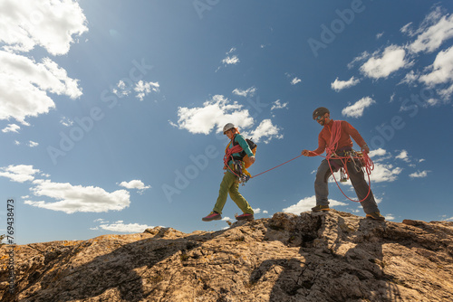 Two people are climbing a mountain, one of them is wearing a red jacket. The sky is blue and there are clouds in the background