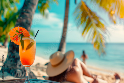 A cocktail in a glass stands on a table near a sun lounger on the beach under the shade of palm trees and a woman in a hat lies on the sun lounger