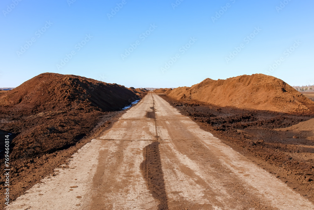 Straight long concrete path through a peat bog with large peat hills and blue sky in the background