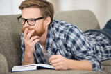 young hipster guy wearing eyeglasses reading a book on sofa