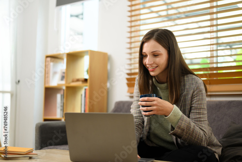 Smiling adult woman drinking coffee and working with laptop in living room