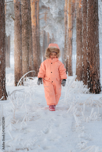 A little girl of 4-6 years old in a pink jumpsuit walks in the middle of a winter pine park, vertical photo