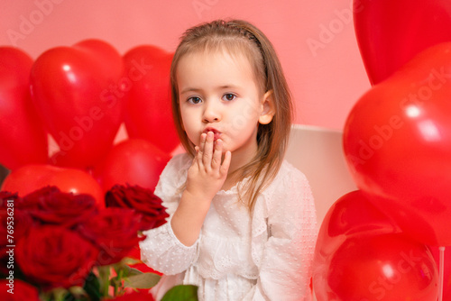 A beautiful baby girl in a white dress blows a kiss while looking at the camera, posing on a pink background.