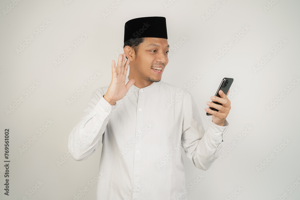 Happy smiling Asian Muslim man standing with Eid Mubarak greeting gesture and welcoming Ramadan video call via mobile phone - obrazy, fototapety, plakaty 
