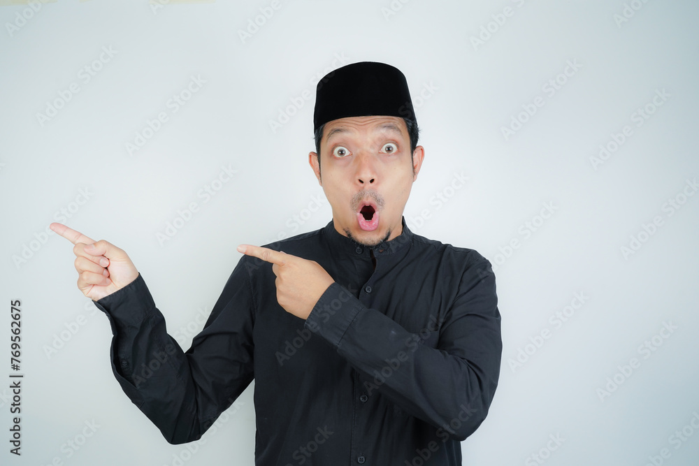 Wow face shocked expression Asian Muslim man pointing hand finger at empty space on isolated background - obrazy, fototapety, plakaty 