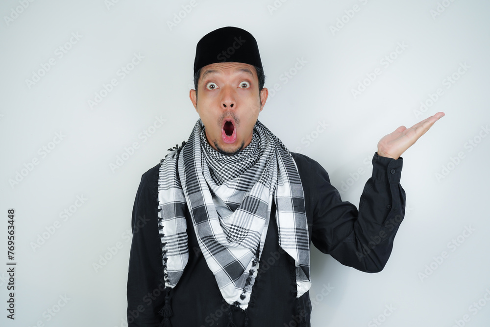 Wow face shocked expression Asian Muslim man wearing Arab turban sorban pointing hand finger at empty space on isolated background - obrazy, fototapety, plakaty 
