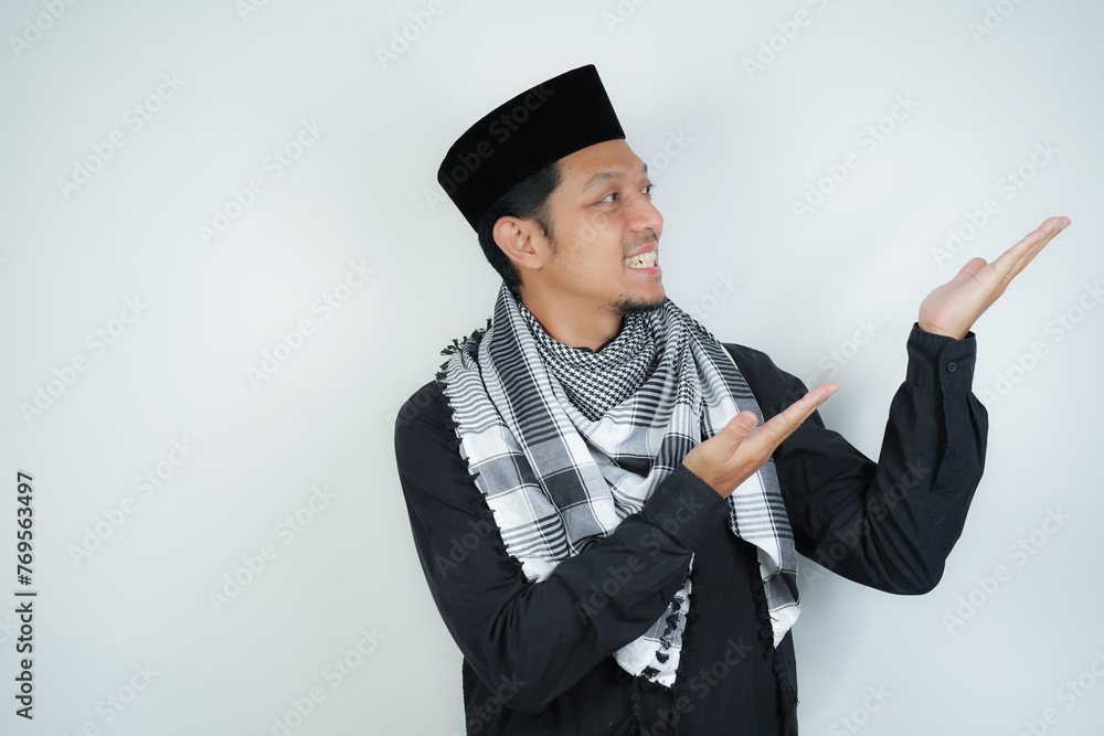 Cheerful handsome Asian Muslim man wearing Arab turban sorban pointing hand finger at empty space for advertising on isolated background - obrazy, fototapety, plakaty 