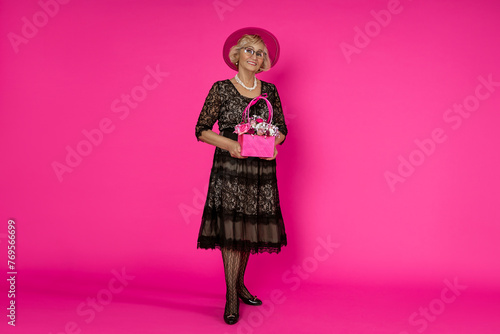 Beautiful happy senior woman in elegant black dress holding a bouquet with flowers on a pink background in the studio.Mother's Day and Grandmother's Day concept.