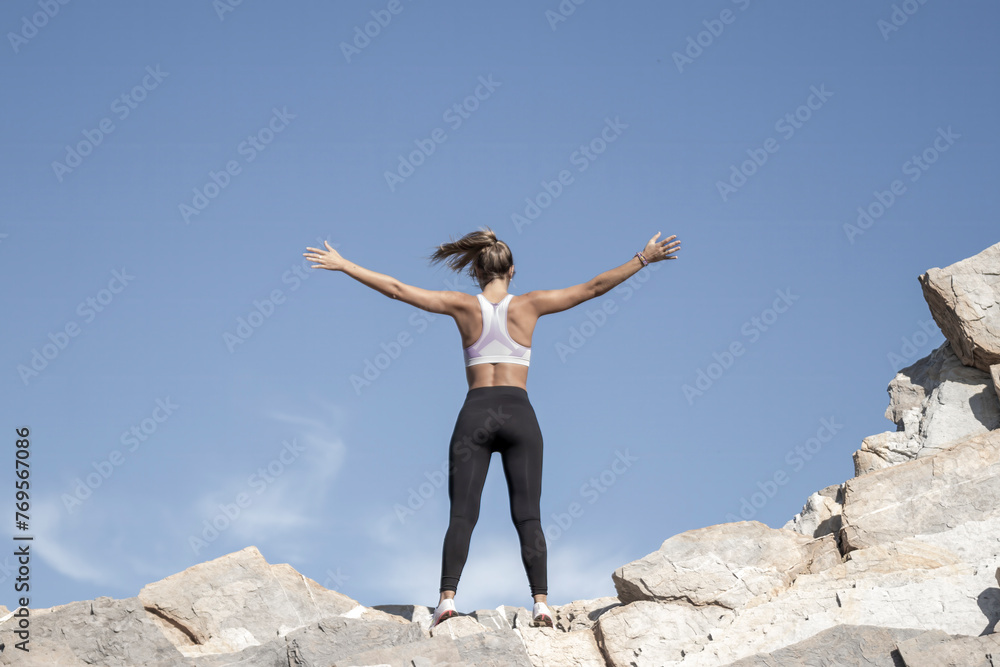 A woman stands triumphantly atop rocky terrain, arms outstretched, under a clear blue sky, embodying freedom and achievement A woman stands triumphantly atop rocky terrain, arms outstretched, under a clear blue sky, embodying freedom and achievement