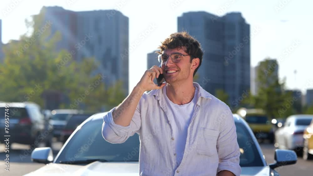 Happy young man in glasses leaning on the car while talking on the smartphone, standing outdoors. Transport, technology and lifestyle concept. Slow motion Happy young man in glasses leaning on the car while talking on the smartphone, standing outdoors. Transport, technology and lifestyle concept. Slow motion