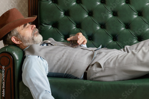 An elderly, tipsy man, half-asleep, leisurely drinks wine while lying on the sofa.
