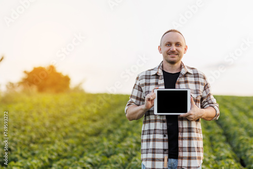 Male farmer shows digital tablet with empty black screen on green soybeans field. Smart farming technology and organic agriculture concept. Quality control growth and development soya plants