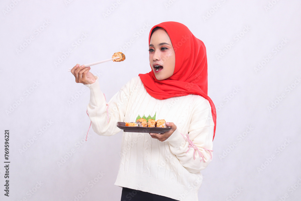 Asian girl wearing a hijab surprised candid holding chopsticks eating and carrying a plate containing sushi (Japanese food) while offering. Beautiful Muslim women use culinary, food and lifestyle Asian girl wearing a hijab surprised candid holding chopsticks eating and carrying a plate containing sushi (Japanese food) while offering. Beautiful Muslim women use culinary, food and lifestyle