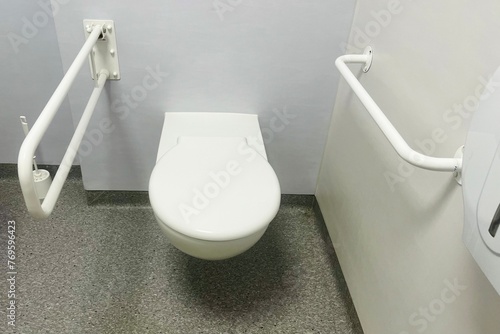 white toilet with metal grab bars in a toilet in a hospital with gray walls. front view