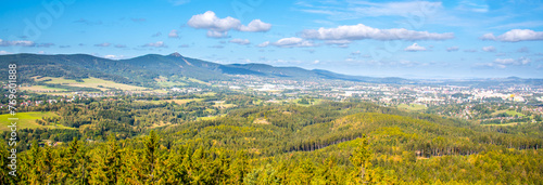 A sweeping vista of Liberec city nestled between rolling hills and Jested ridge under a blue sky dotted with clouds.