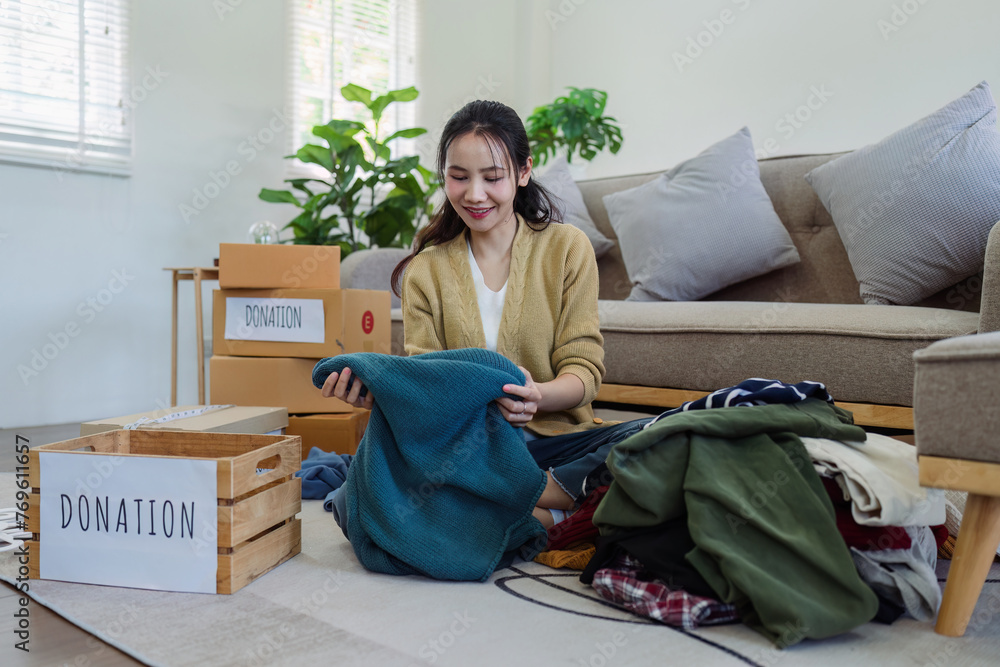 Obraz premium Asian young woman packing clothes at home, putting on stuff into donate box with second hand clothes. Help poor people. Donation concept