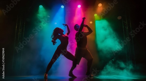 Silhouettes of two dancers in motion on stage with vibrant lighting and smoke effects. © cherezoff