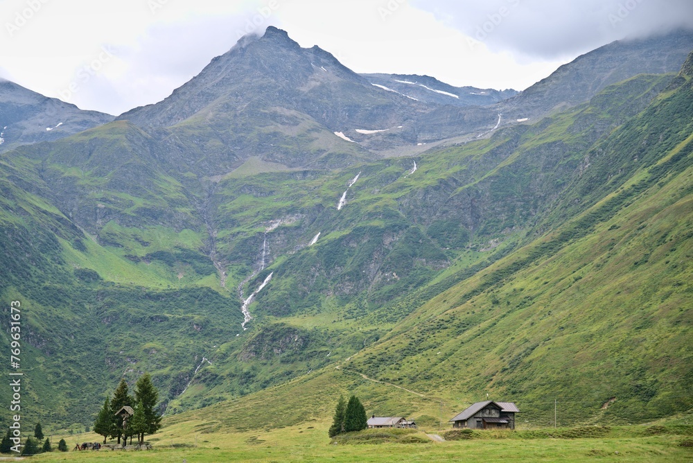 Wooden lodge with a high mountain and waterfall behind, lush green grass in Austrian Alps