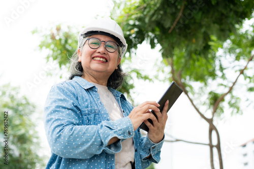 Happy elderly Asian professional engineer wears a helmet and checks the blueprint on a tablet on the construction site after the contractor and architect finish renovation the house and building