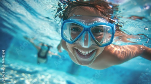 A joyful underwater scene with a person wearing blue goggles smiling broadly and swimming in clear blue water with another swimmer in the background.