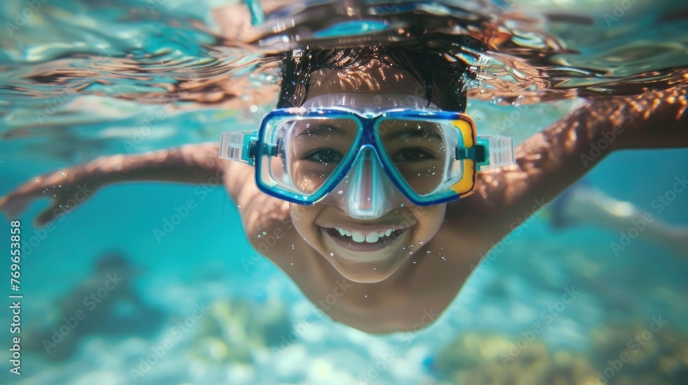 Young boy joyfully swimming underwater with goggles smiling at the camera surrou nded by blue water and aquatic life.
