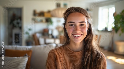 A young woman with a radiant smile wearing a cozy orange sweater seated in a warmly lit inviting living room filled with plants and natural light.