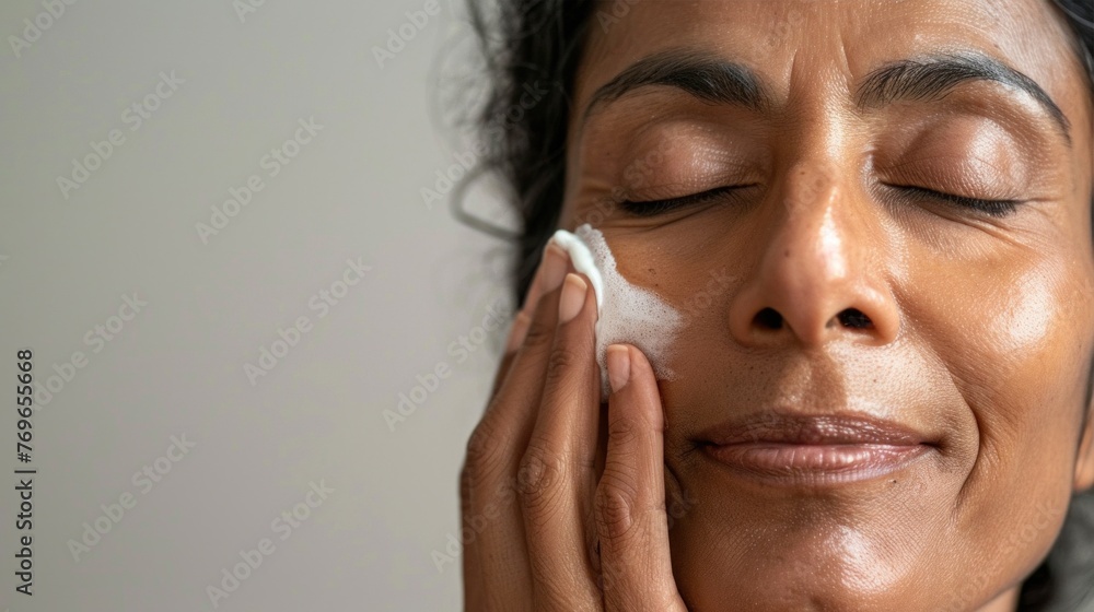 A woman with closed eyes smiling gently massaging her face with a white substance possibly a skincare product in a serene and peaceful setting. A woman with closed eyes smiling gently massaging her face with a white substance possibly a skincare product in a serene and peaceful setting.