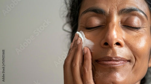 A woman with closed eyes smiling gently massaging her face with a white substance possibly a skincare product in a serene and peaceful setting.