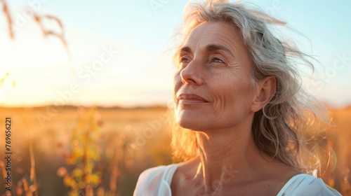 A serene sunset scene with a woman in a white top her hair blowing in the wind looking up at the sky with a peaceful expression.