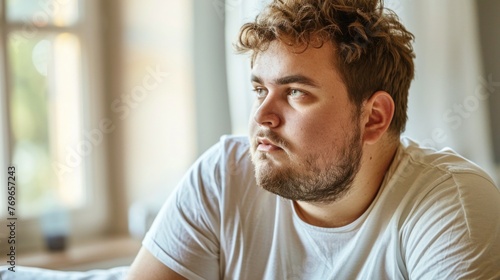 A man with a beard and curly hair wearing a white t-shirt sitting by a window with a thoughtful expression. © iuricazac