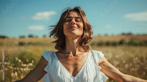 A woman in a white dress with her eyes closed standing in a field of tall grass smiling and enjoying the moment with a clear blue sky in the background. © iuricazac