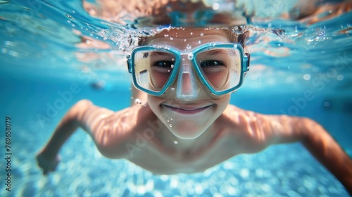 A young boy wearing blue goggles smiling underwater with bubbles around his face in a swimming pool. © iuricazac