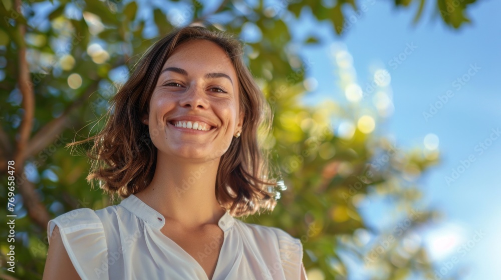 Smiling woman with brown hair and white blouse standing in front of blurred trees and blue sky. Smiling woman with brown hair and white blouse standing in front of blurred trees and blue sky.