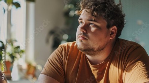 A man with curly hair and a beard wearing a brown t-shirt sitting in a room with plants looking contemplative.