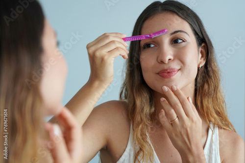 Dermaplaning. Beautiful young woman shaving her eyebrows by razor on the mirror at home. Facial hair removal. photo