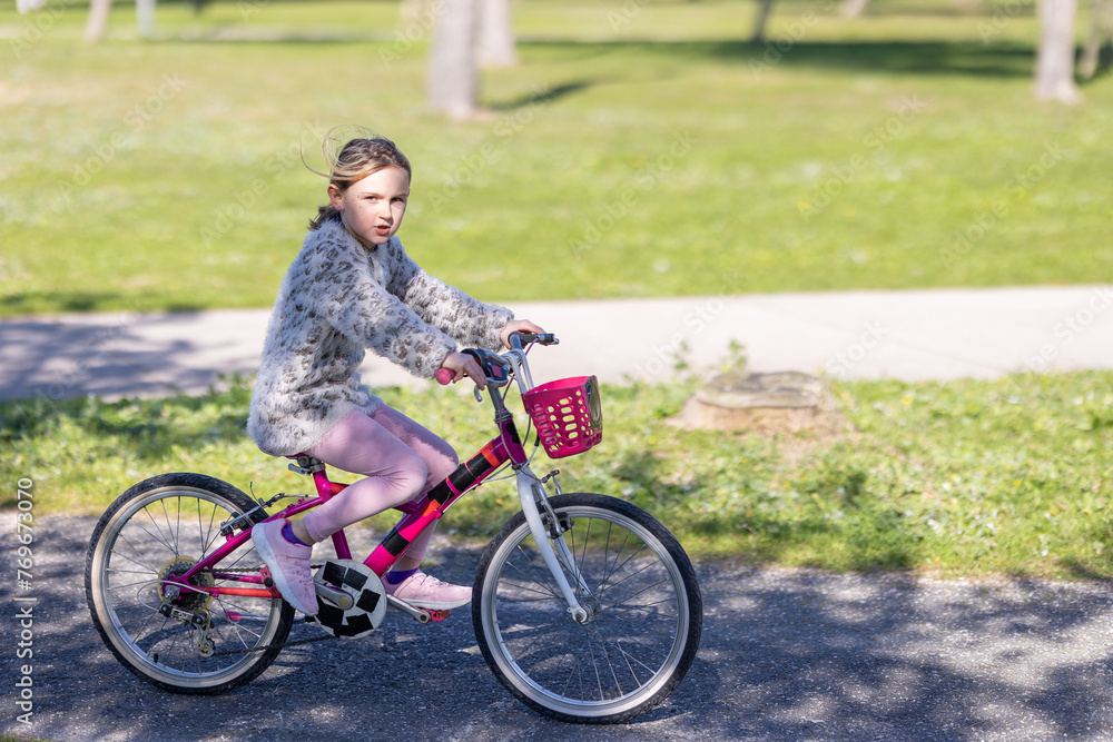 A young girl is riding a pink bicycle with a basket on the front - obrazy, fototapety, plakaty 