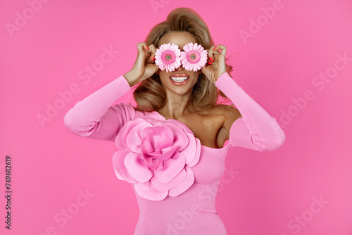 Horizontal shot of pretty woman has blonde hair holds two gerbera daisy in front of eyes and smiles broadly, has fun. Fashionable lady enjoys spring time, pleasant fragrance. Florist with flowers