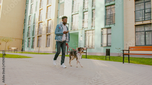 Pet owner with smartphone in hands keeping dog on leash while walking on house surrounding area. African american man wearing casual clothes checking messages during walk.