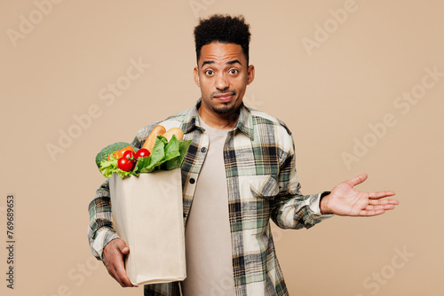 Young man wear grey shirt hold paper bag for takeaway mock up with food products spread hands shrugging shoulders isolated on plain pastel beige background. Delivery service from shop or restaurant.