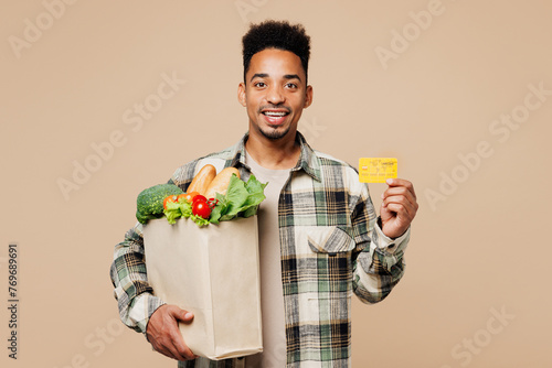 Young smiling man wears grey shirt hold paper bag for takeaway mock up with food products, credit bank card isolated on plain pastel light beige background. Delivery service from shop or restaurant.