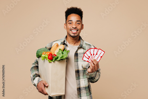 Young fun man wear grey shirt hold paper bag for takeaway mock up with food products, show many coupon cards isolated on plain pastel light beige background. Delivery service from shop or restaurant.