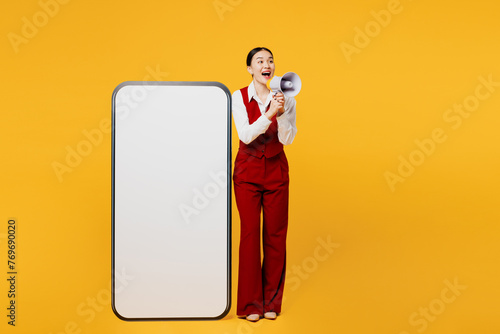 Full body young lawyer employee business woman of Asian ethnicity wear formal red vest shirt work at office big blank screen mobile cell phone scream in megaphone isolated on plain yellow background.