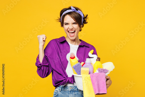 Young excited woman wear purple shirt hold basin with detergent bottles do housework tidy up doing winner gesture celebrate isolated on plain yellow background studio portrait. Housekeeping concept.