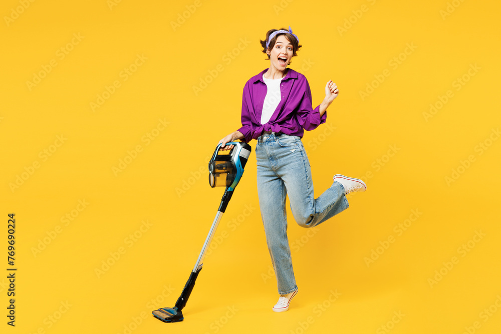 Full body happy young woman wear purple shirt casual clothes do housework tidy up hold vacuum cleaner raise up leg look camera isolated on plain yellow background studio portrait Housekeeping concept