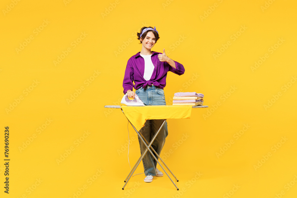 Full body young woman she wear purple shirt casual clothes do housework tidy up ironing clean clothes on board show thumb up isolated on plain yellow background studio portrait. Housekeeping concept. - obrazy, fototapety, plakaty 