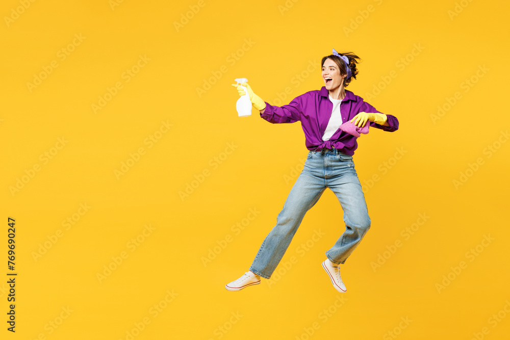 Full body fun young woman wears purple shirt rubber gloves do housework tidy up hold in hand rag use spray bottle jump high isolated on plain yellow background studio portrait. Housekeeping concept.